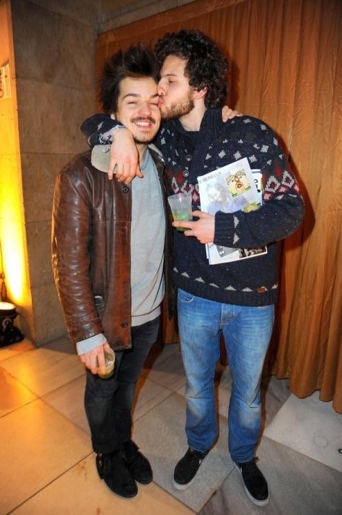 Philipp in a black sweater and blue jeans kissing his friend on his forehead.